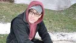 A young man, glasses, baseball hat and a hoodie, sitting outside. 