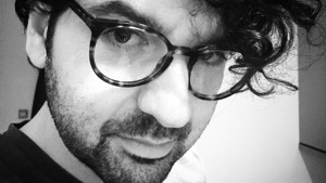 Close up black and white photo of Paolo Santorio, Professor of Philosophy, leaning forward into the camera, with a serious expression, black ringlets of hair tumbling over his round glasses.