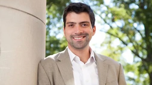 Portrait of a man in a beige sportscoat, from the chest up, standing outside, with a confident smile.