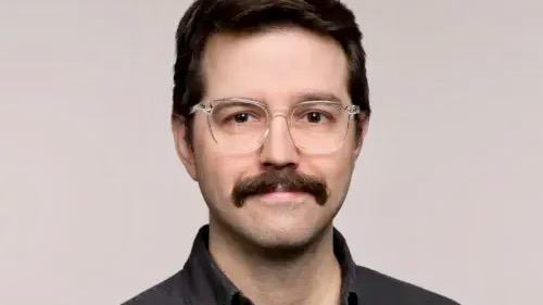 A young man with clear-framed glasses and a moustache.