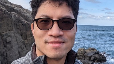 A young man wearing glasses, standing on a rocky coastline.