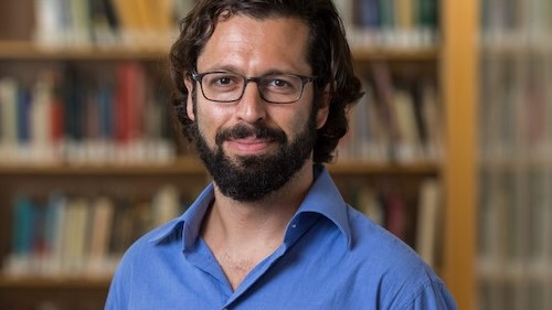 A bearded young man in a blue shirt and glasses, standing in front of a bookcase.