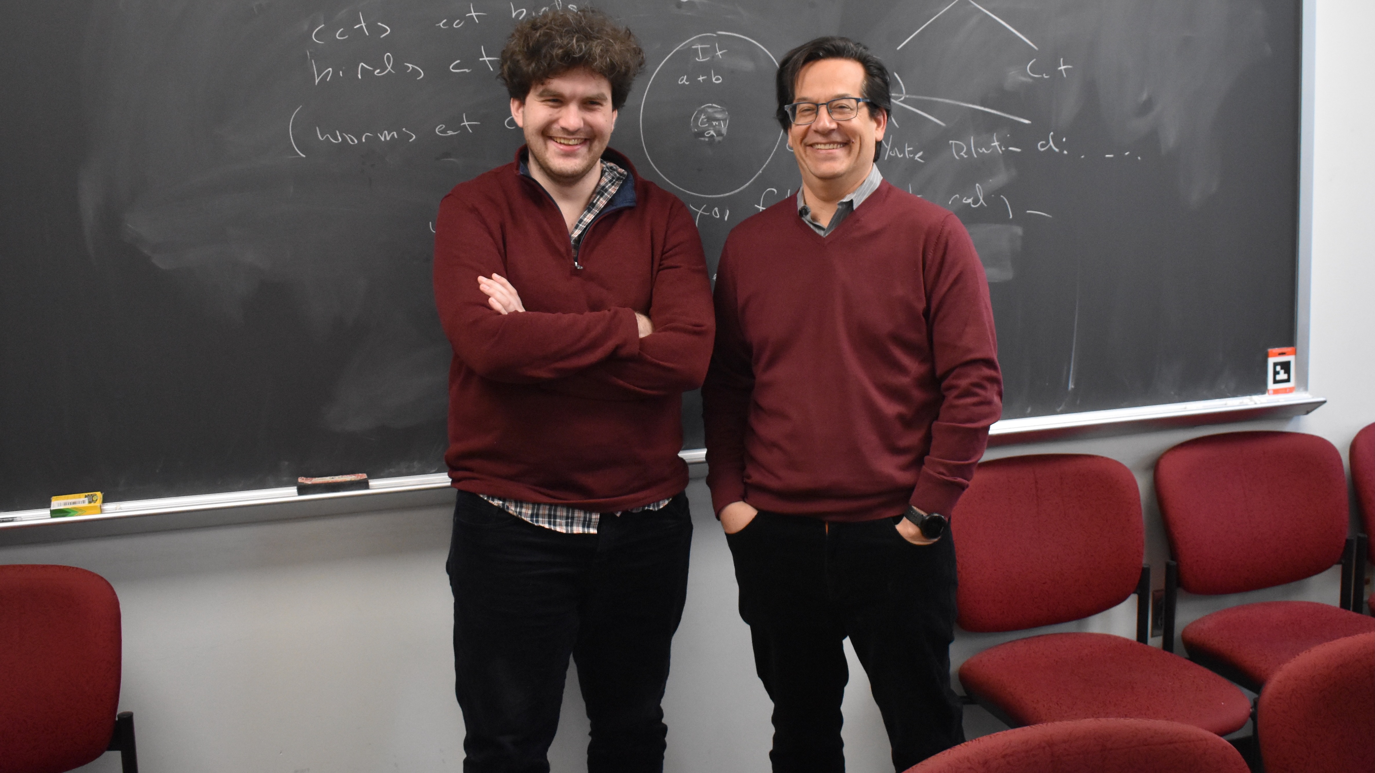 Two men standing next to each other, in front of a blackboard, both wearing burgundy v-neck sweaters, and dark blue corduroy pants with a somewhat velvety surface.