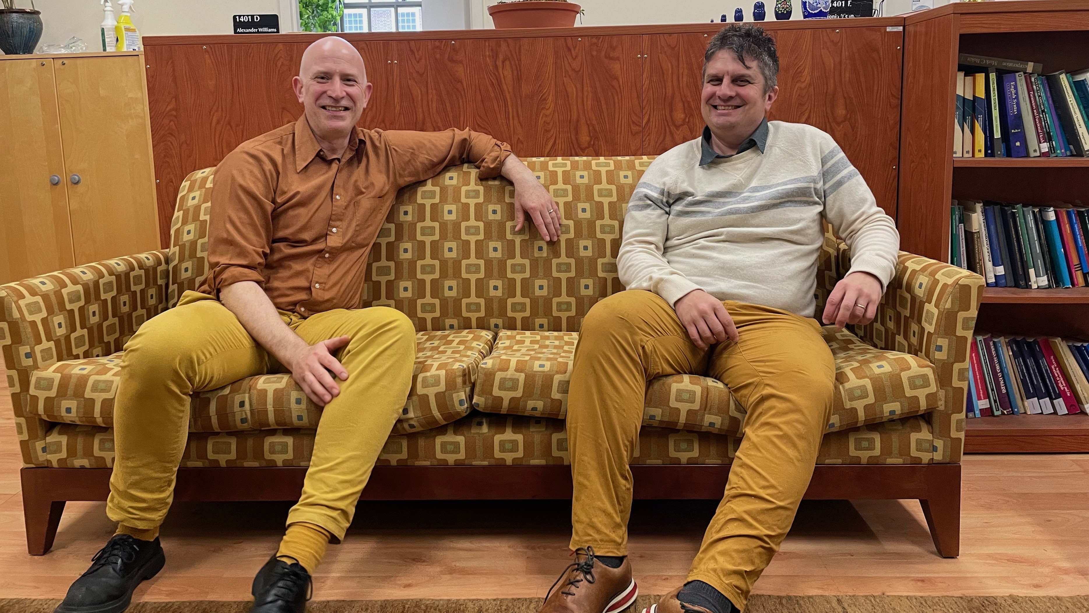 Two middle-aged men, wearing harvest gold and amber brown clothing, sitting on a sofa with a mod pattern in harvest gold and amber brown.