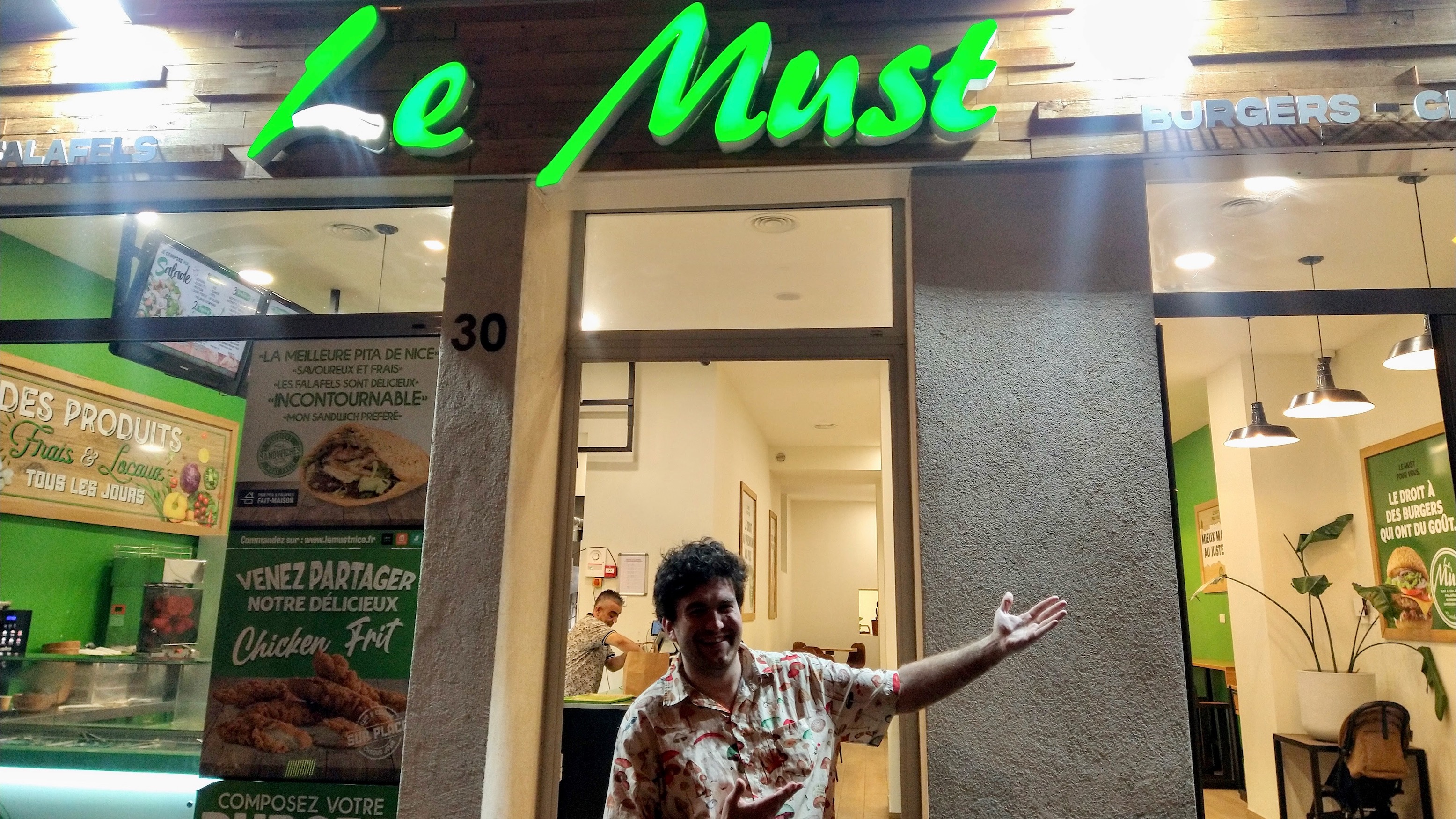 A man standing in front of a hamburger restaurant in France, called "Le Must", gesturing towards the sign.