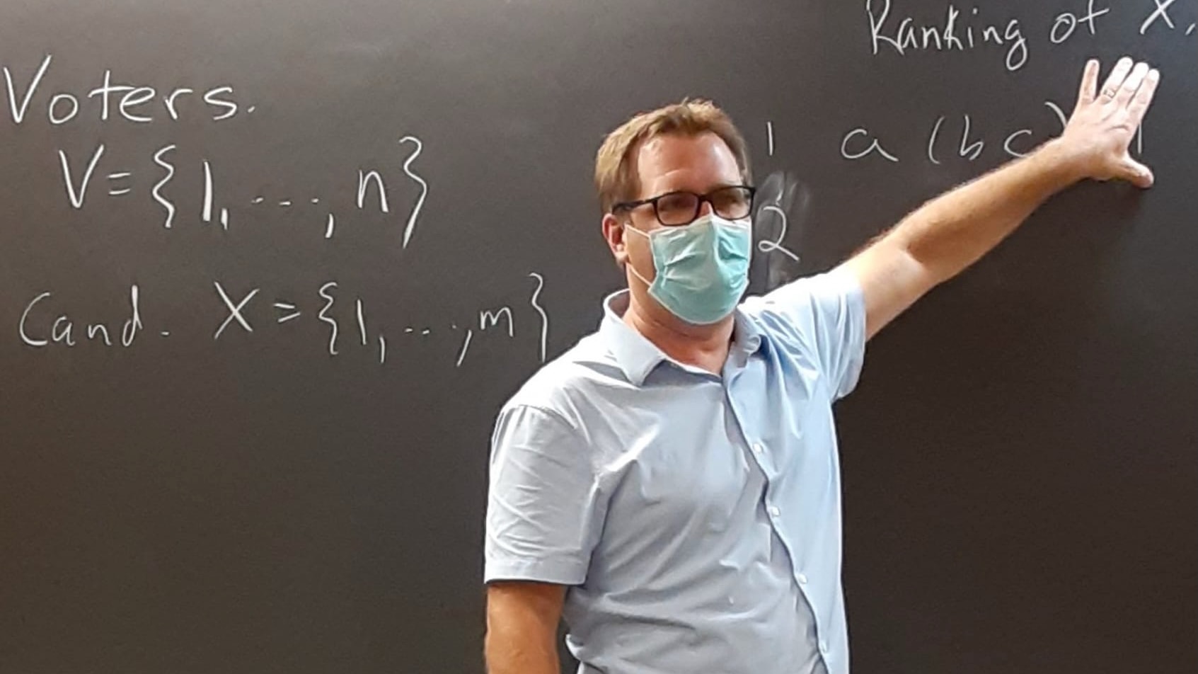 A man in a covid mask, standing in front of a chalkboard with some formulas written on it.