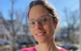 Portrait of a woman wearing glasses, outside, with a blue sky and bare trees behind her.