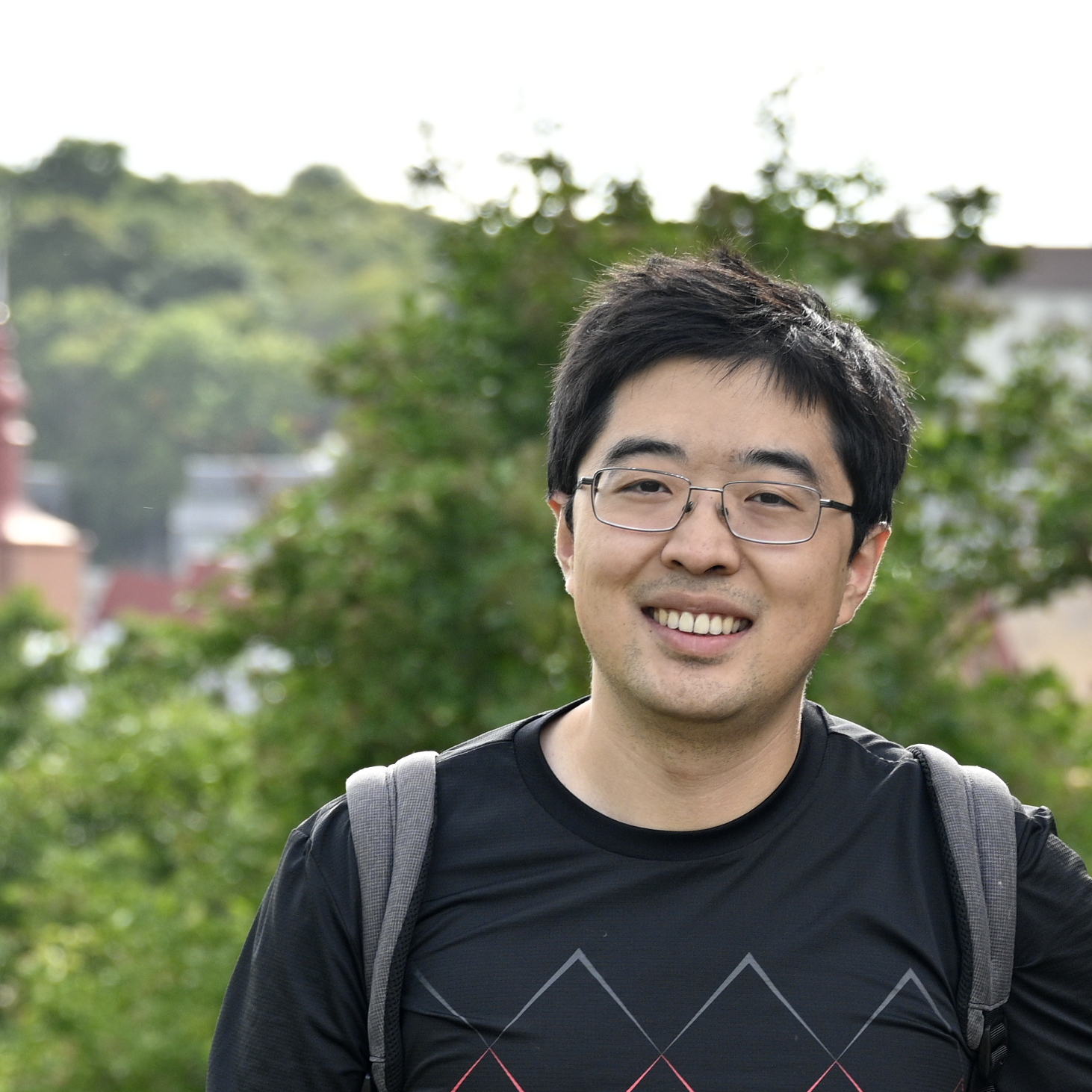 A young man standing outside, wearing a rucksack, small trees and a village in the background.