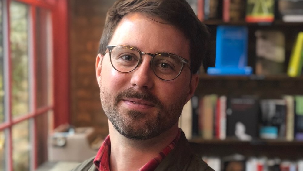A man with a short beard and glasses, sitting in his home study, head slightly tilted, with a gentle smile.