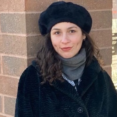 A young woman in a winter beret and coat, standing in front of a brick column, smiling faintly.