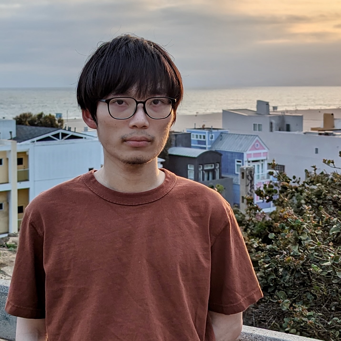 A young man standing on a hill in a southern California town, with the sun setting over the Pacific Ocean in the background.