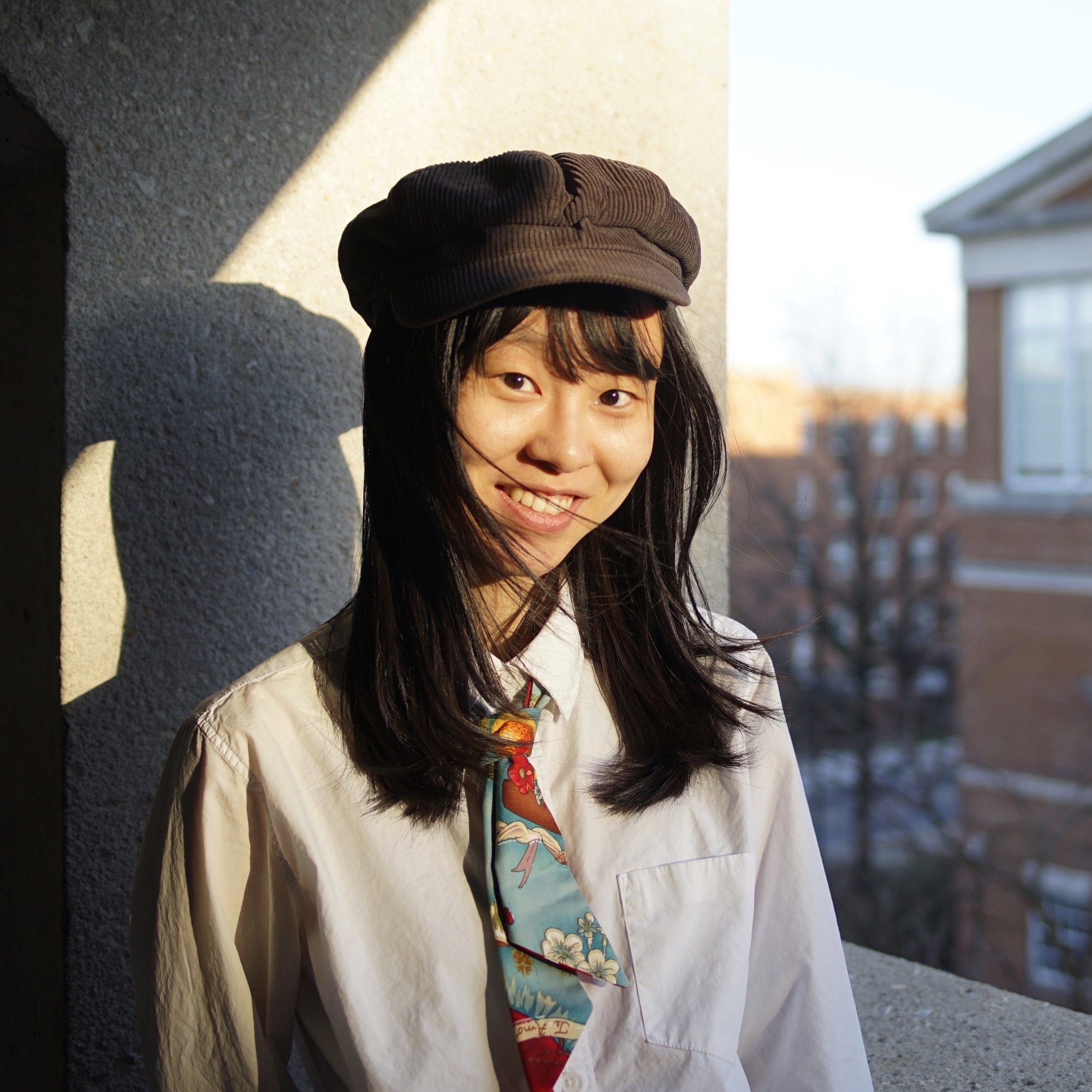 A young woman wearing a cap, standing on a city balcony, smiling.