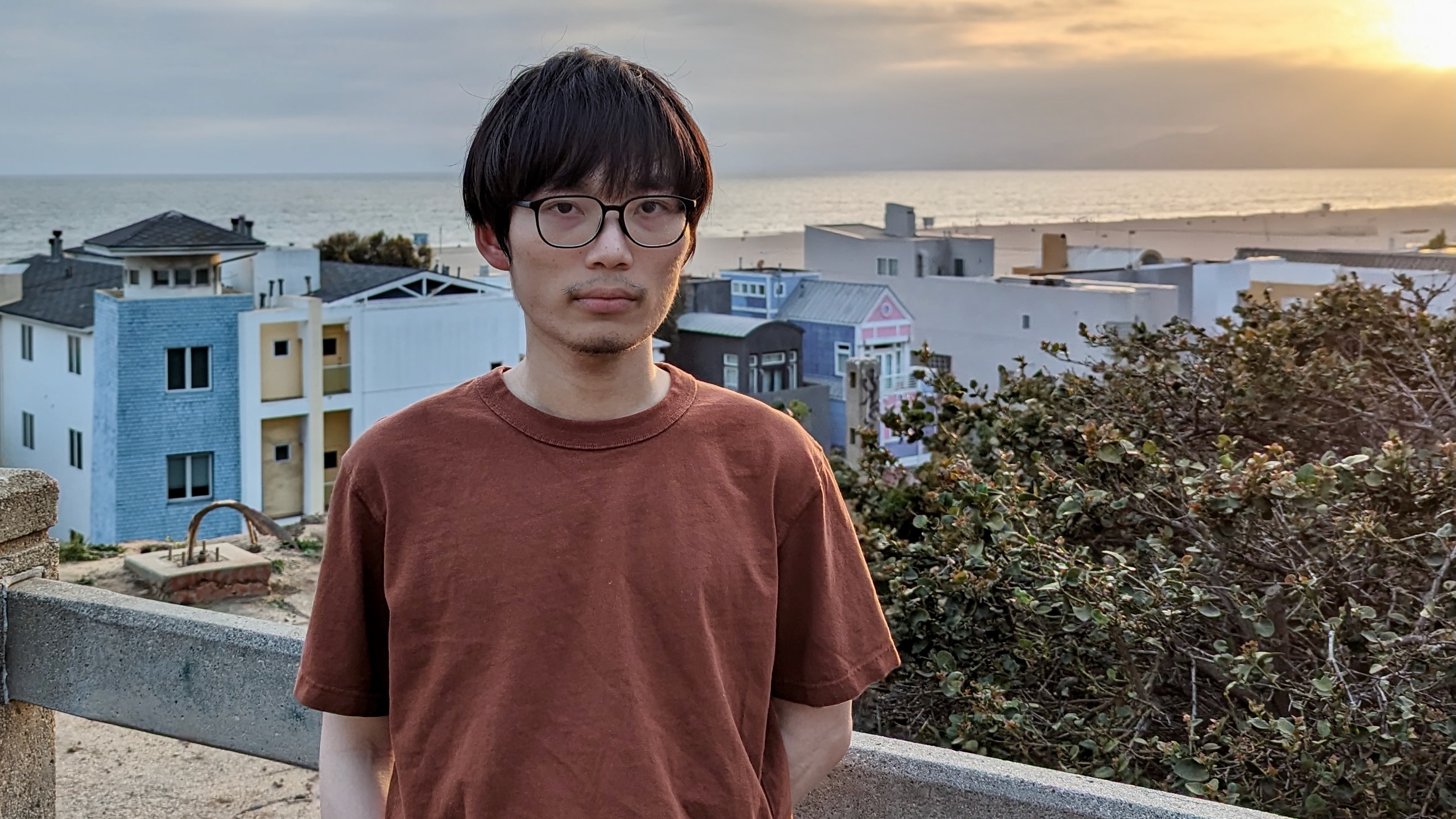 A young man standing on a hill in a southern California town, with the sun setting over the Pacific Ocean behind him.