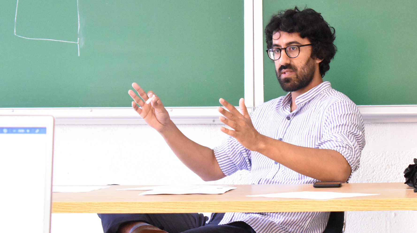 A bearded man sitting in front of a green chalkboard, with his hands up as if to measure a 2ft length, teaching a class.