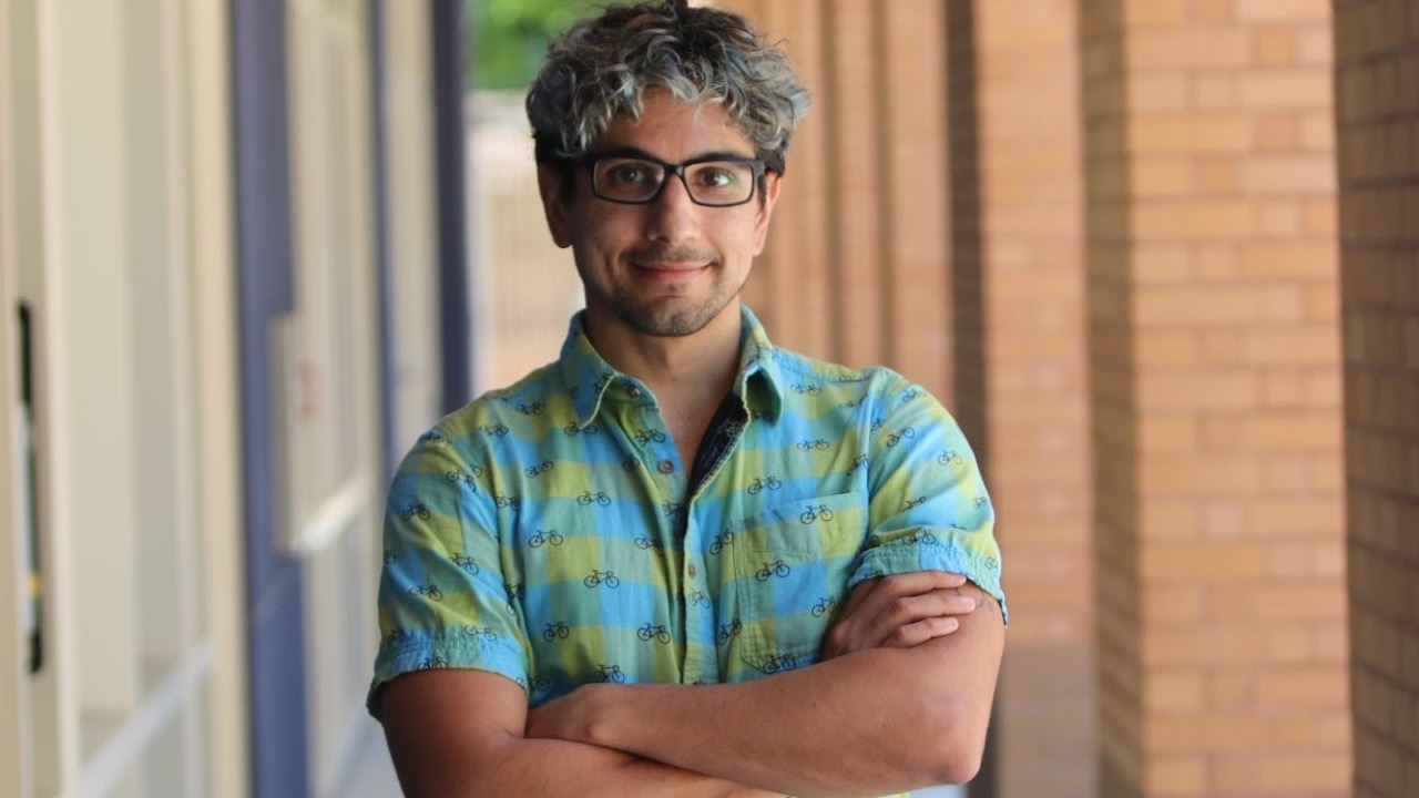 A man standing outside, under a portico, wearing a short-sleeve blue-green shirt.