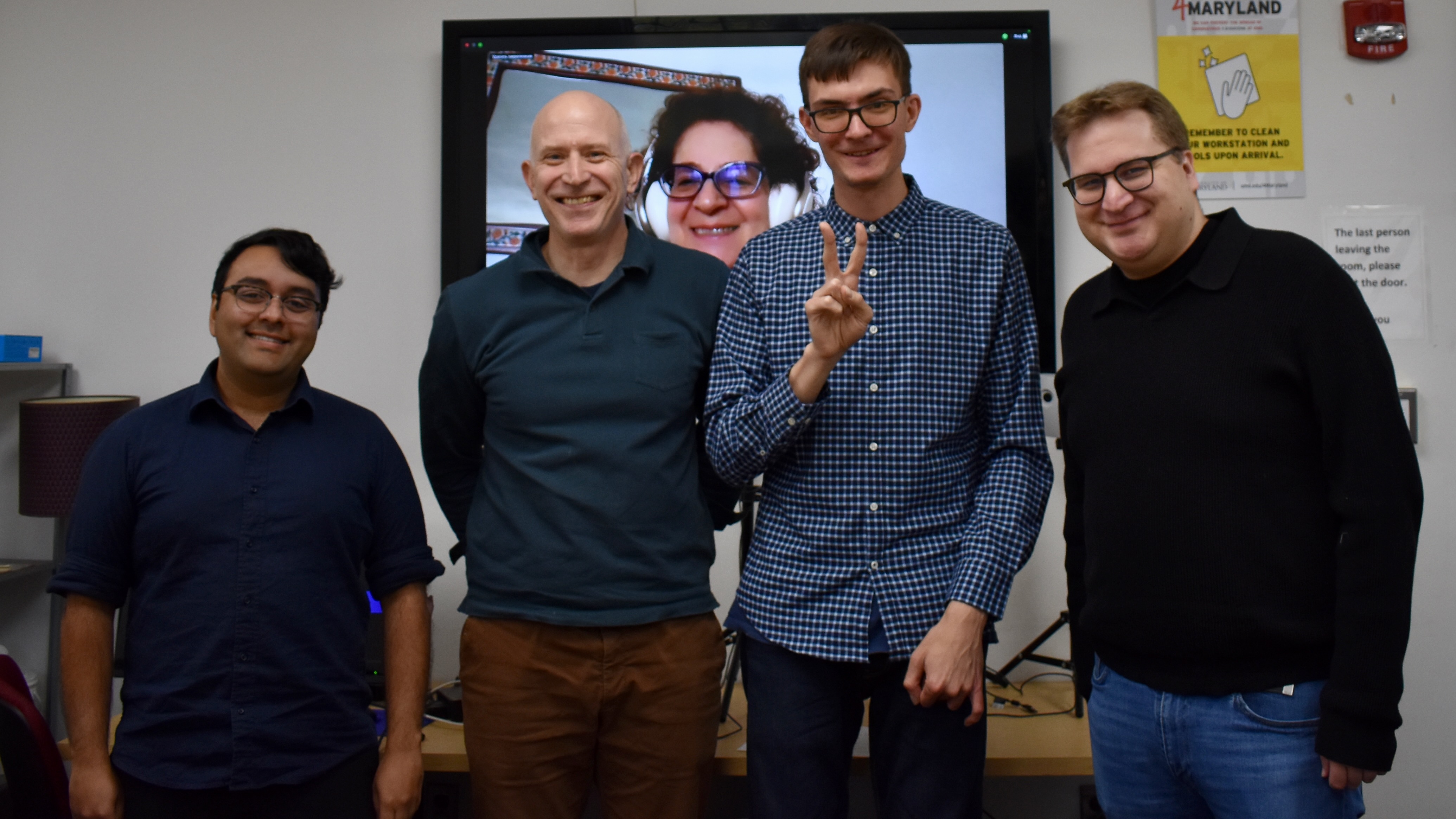 Four men standing in front of a large TV screen, smiling in a ceremonial pose, with a woman's head on the screen behind them, smiling together with them.